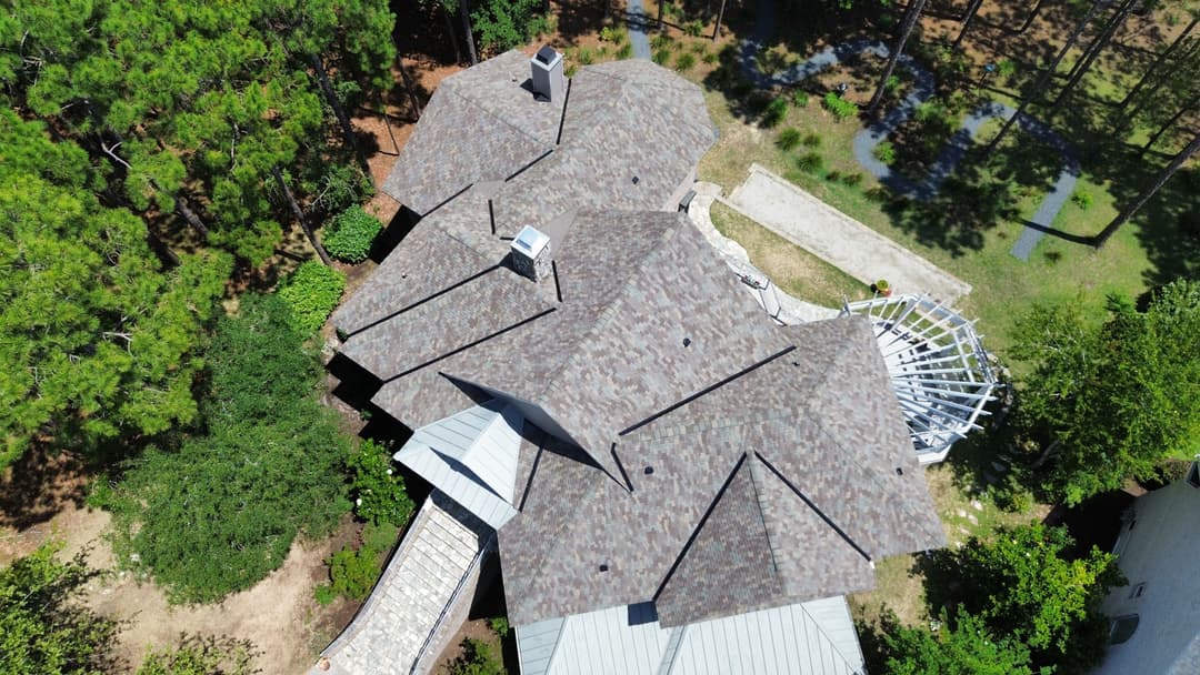 Aerial view of a house with a multi-gable roof surrounded by trees and a patio area.