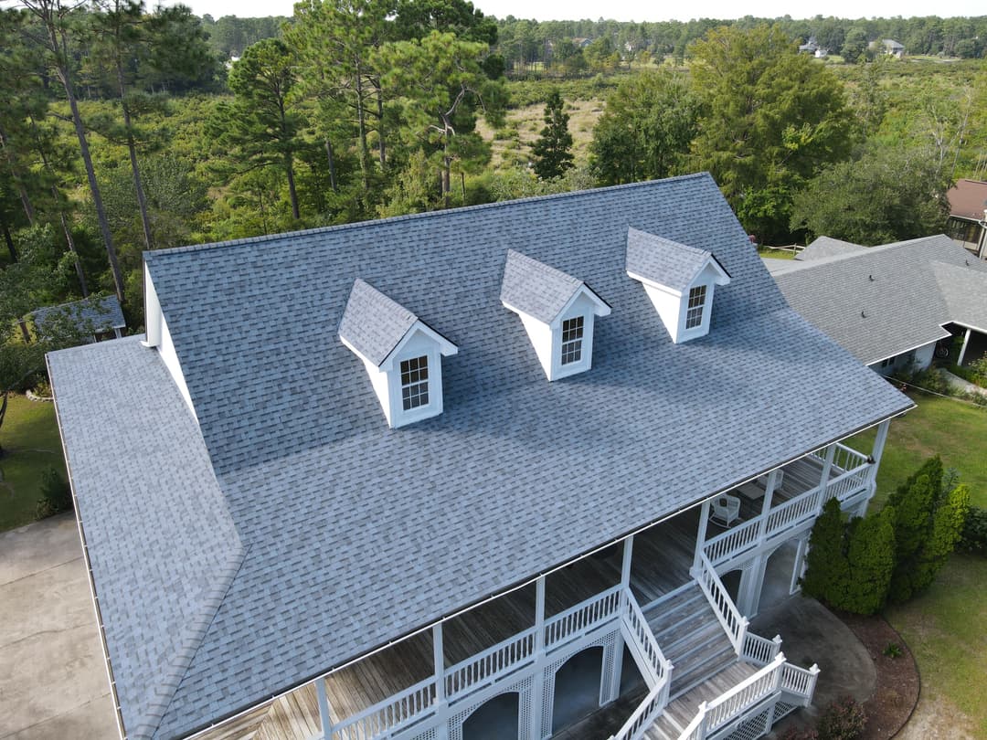 Aerial view of a gray shingle roof with three gable dormers on a house surrounded by trees.