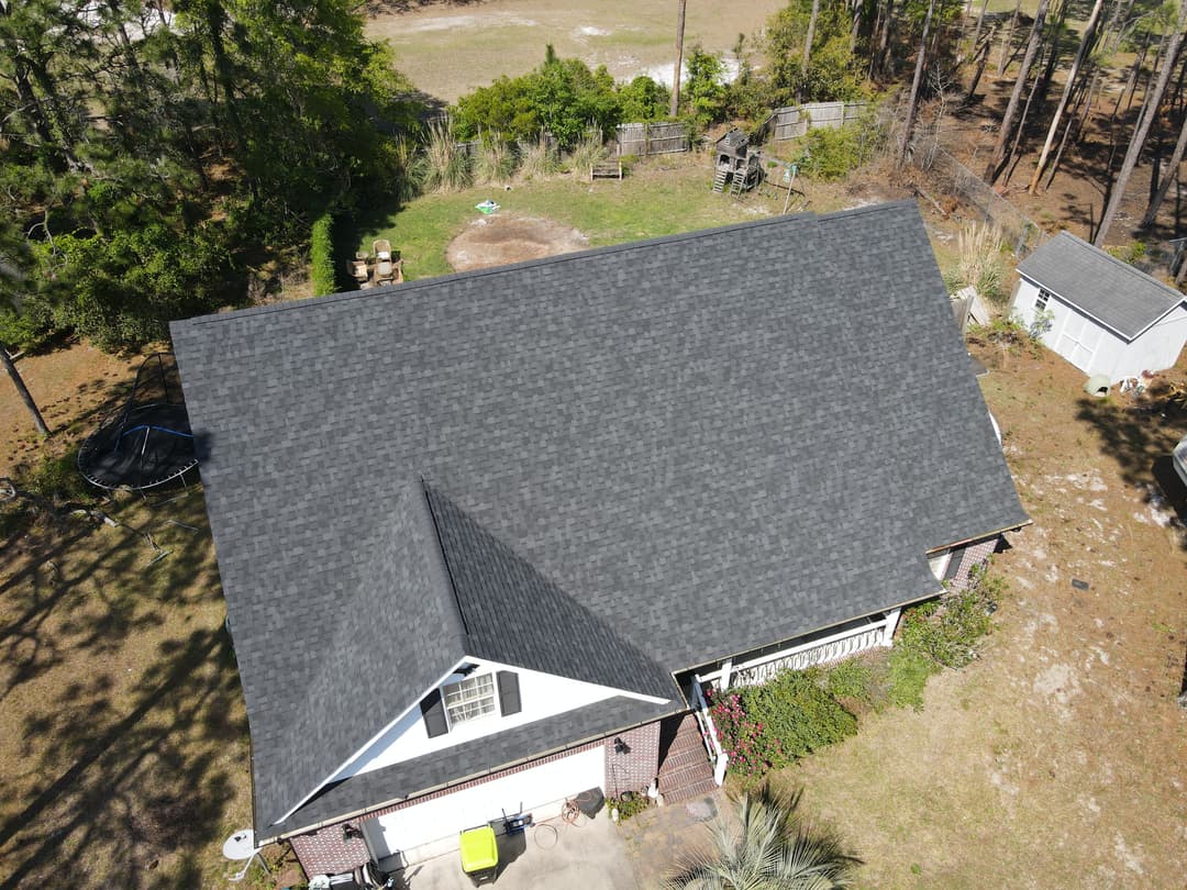 Aerial view of a residential house with a black shingle roof and surrounding trees.