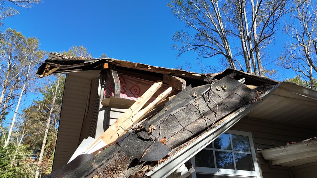 damaged roof with missing shingles and exposed wood against a clear blue sky