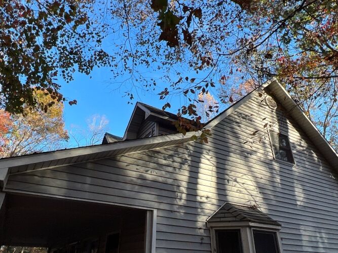 A house with light-colored siding, an attached carport, and a pitched roof, surrounded by trees with autumn foliage under a clear blue sky.