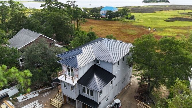 A multi-story house with a dark metal roof is under construction near a marshy area with water and trees in the background. Building materials are scattered around the property.