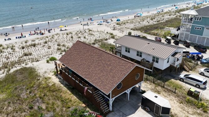 Aerial view of a beach house near sand dunes, with adjacent homes, a trailer, and people on the nearby beach under umbrellas.