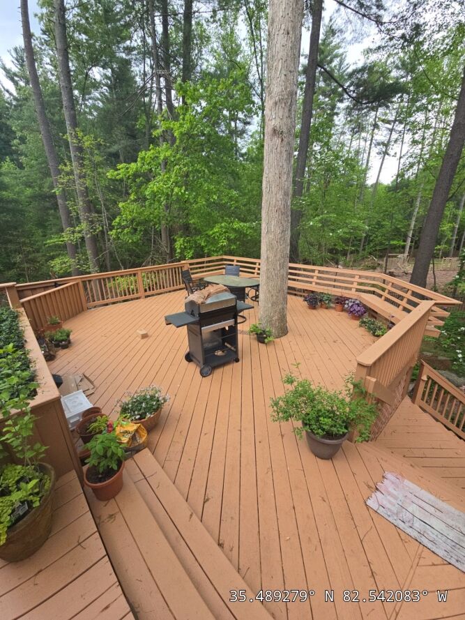 Spacious wooden deck surrounded by trees, featuring a grill, potted plants, chairs, and a tree growing through the deck. GPS coordinates are displayed in the bottom right corner.
