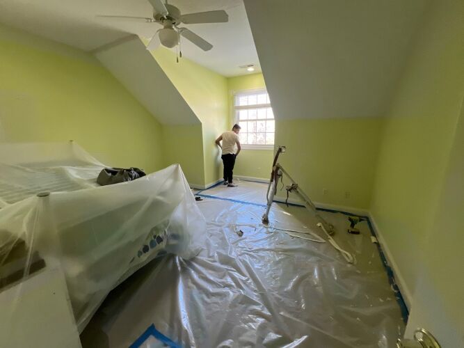 A person stands near a window in a room being painted; furniture is covered with plastic and the floor is protected by a plastic sheet.